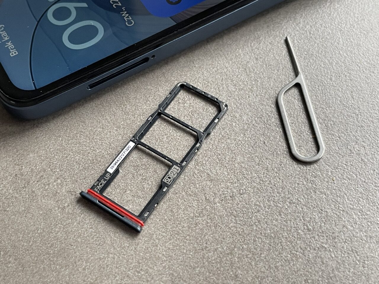 Tacka obok dwóch obiektów: plastikowa tacka SIM z telefonu komórkowego i metalowe narzędzie do wyjmowania tacki SIM, na górze zamazanego ekranu smartfona.