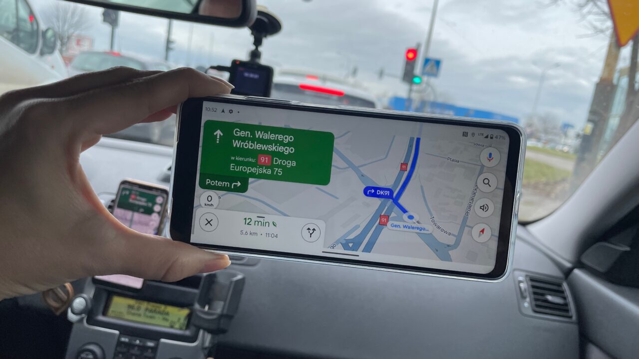 Dłoń trzymająca smartfon z włączoną aplikacją nawigacyjną przedstawiającą mapę i wskazówki dojazdu na trasie, wnętrze samochodu w tle.