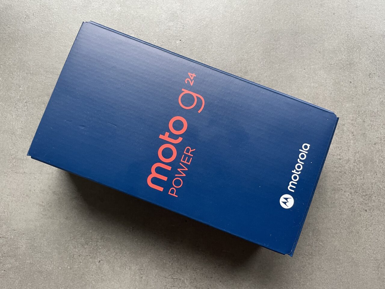 Niebieskie opakowanie smartfona Moto G Power 2021 z logotypem Motorola, położone na szarej powierzchni.