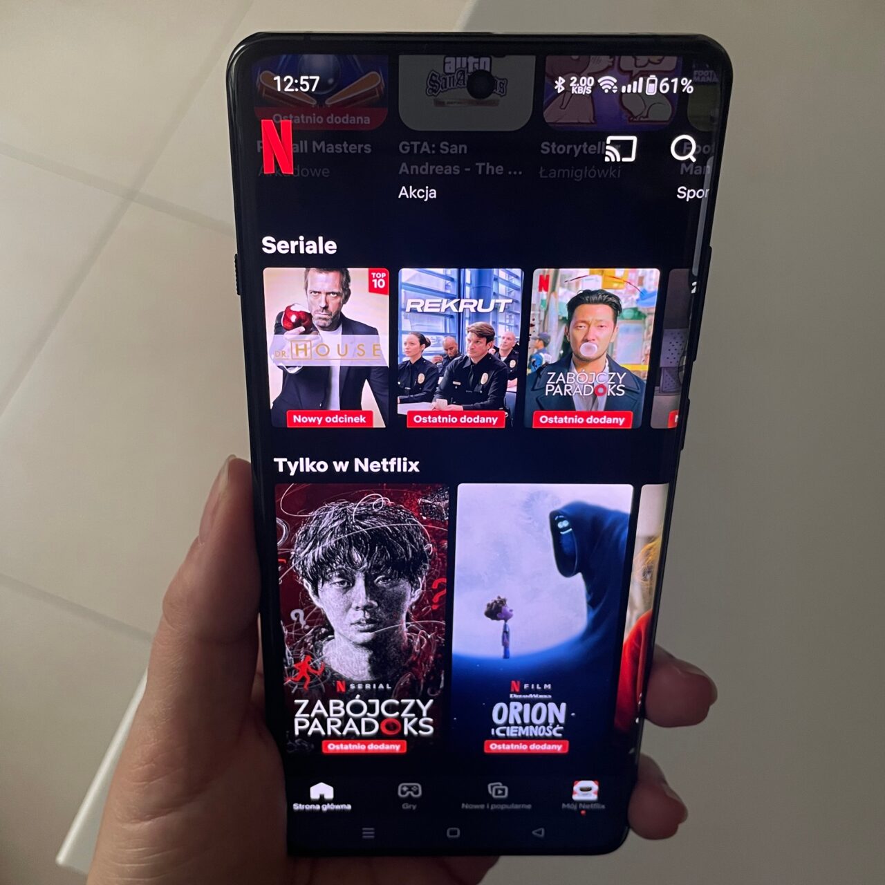Ręka trzymająca smartfon z otwartą aplikacją Netflix wyświetlającą różne tytuły filmów i seriali.