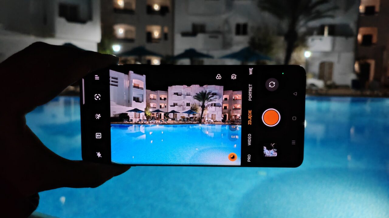 Dłoń trzymająca smartfon, który robi zdjęcie oświetlonego wieczorem budynku przy basenie.