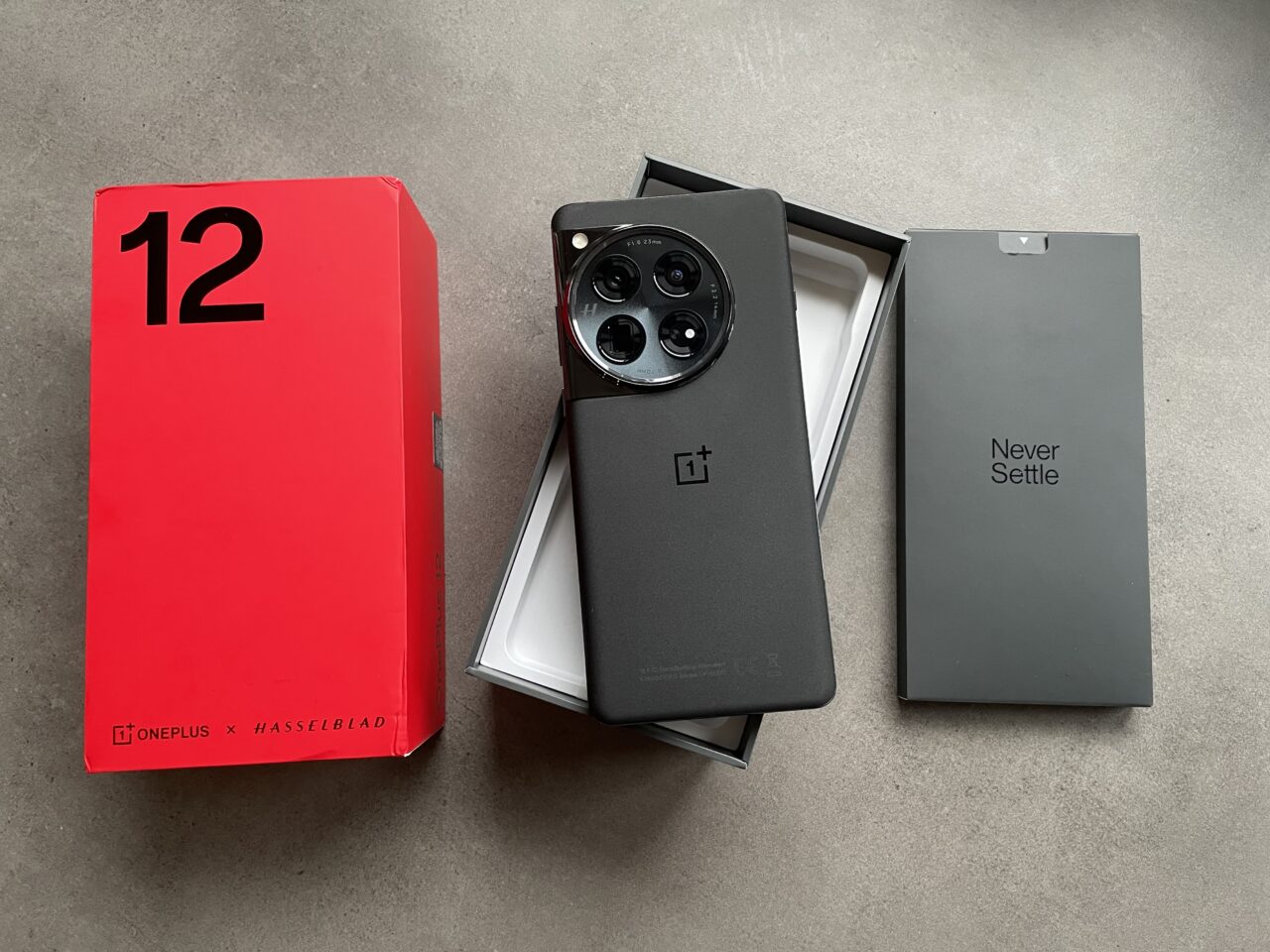 Czerwone pudełko OnePlus z napisem "12" i logotypem Hasselblad, obok otwarte opakowanie z czarnym smartfonem OnePlus i aparatem z trzema obiektywami, oraz szare opakowanie z napisem "Never Settle".