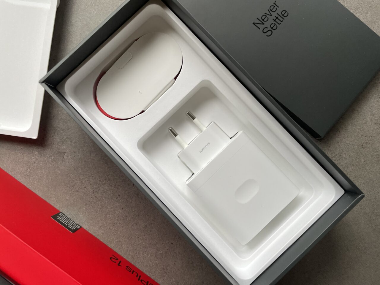 Białe pudełko z akcesoriami elektronicznymi, w tym bielą myszką komputerową z czerwonym akcentem i białą ładowarką sieciową, obok czarnego pudełka z napisem "Never Settle".