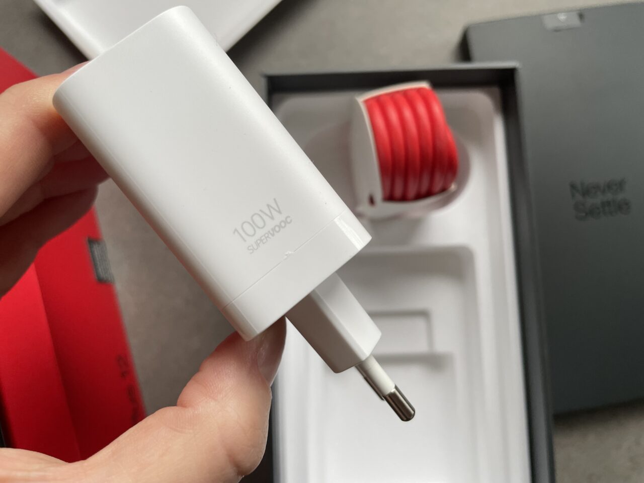 Biała ładowarka sieciowa 100W trzymana w dłoni nad otwartym pudełkiem z czerwonym przewodem USB i ciemnoszarym opakowaniem z napisem "Never Settle".