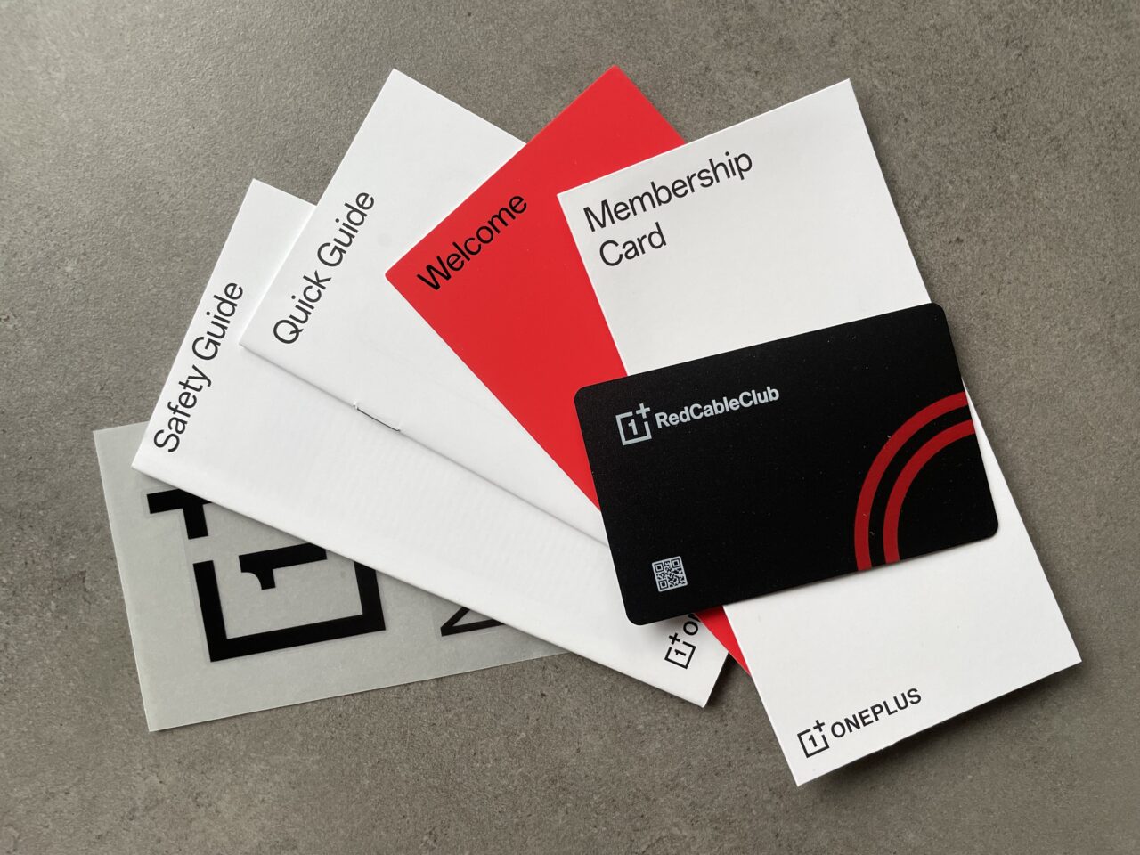 Rozmieszczone na szarym tle materiały powitalne firmy OnePlus, w tym przewodniki oraz czarna karta członkowska Red Cable Club.
