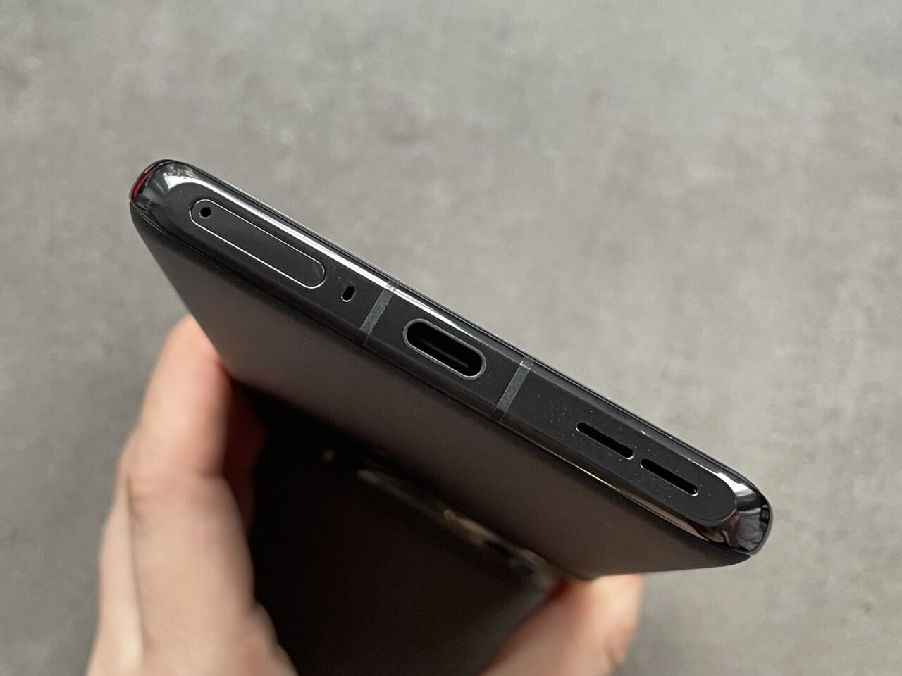 Przytrzymywany w dłoni czarny smartfon z widocznym górnym brzegiem, na którym znajdują się głośnik, port ładowania USB-C, mikrofon i złącze słuchawkowe.