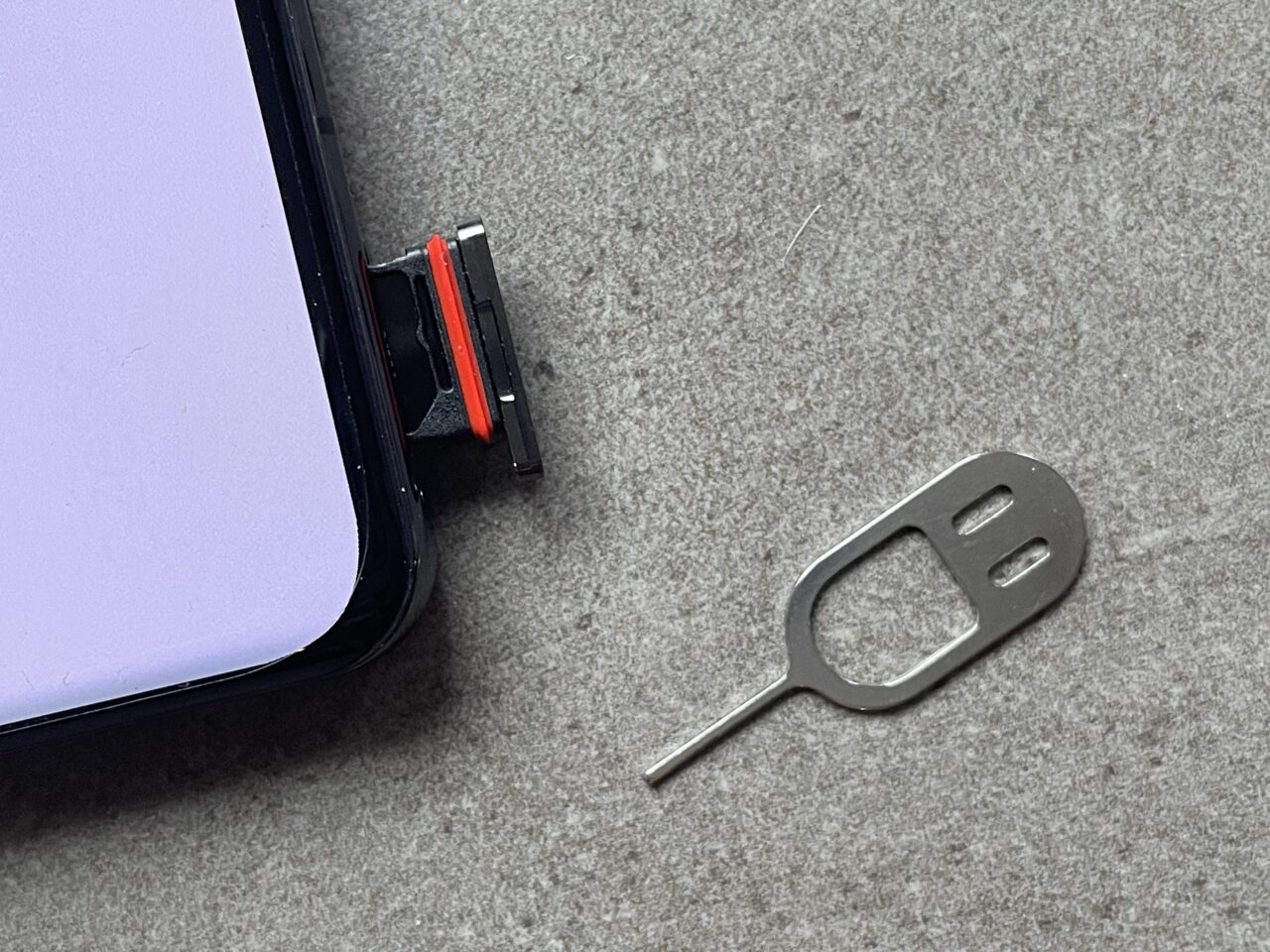 Zdjęcie karty SIM na szarej tkaninie obok metalowego narzędzia do otwierania tacki karty SIM i częściowo widocznego czarnego smartfona z fioletowym ekranem.