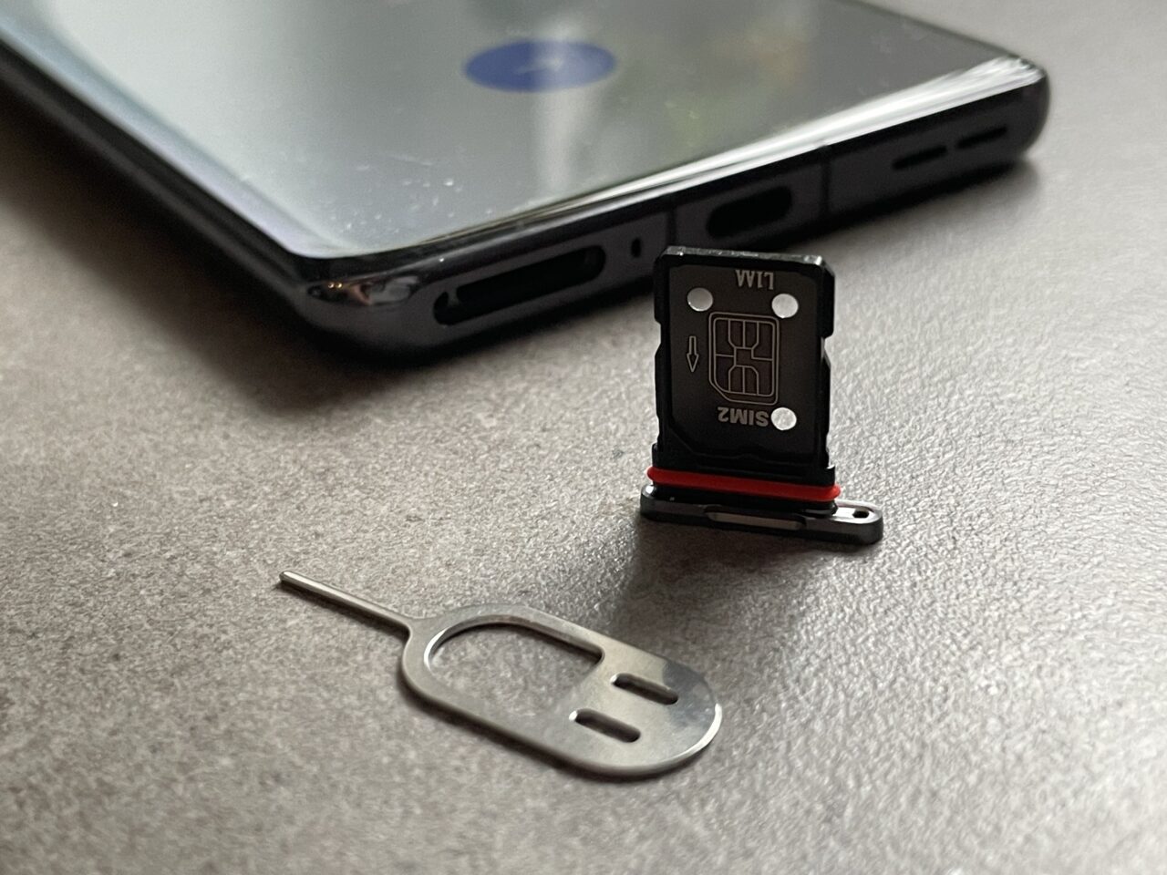 Taca z otwartym gniazdem na karty SIM i microSD obok leżącego narzędzia do wyjmowania tacki z telefonu, na tle ciemnej powierzchni.