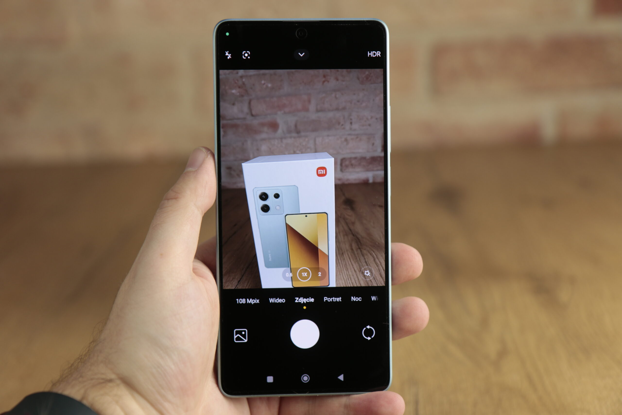 Ręka trzymająca testowany smartfon Redmi Note 13 5G, na którym widać interfejs aparatu fotografującego opakowanie innego smartfona.