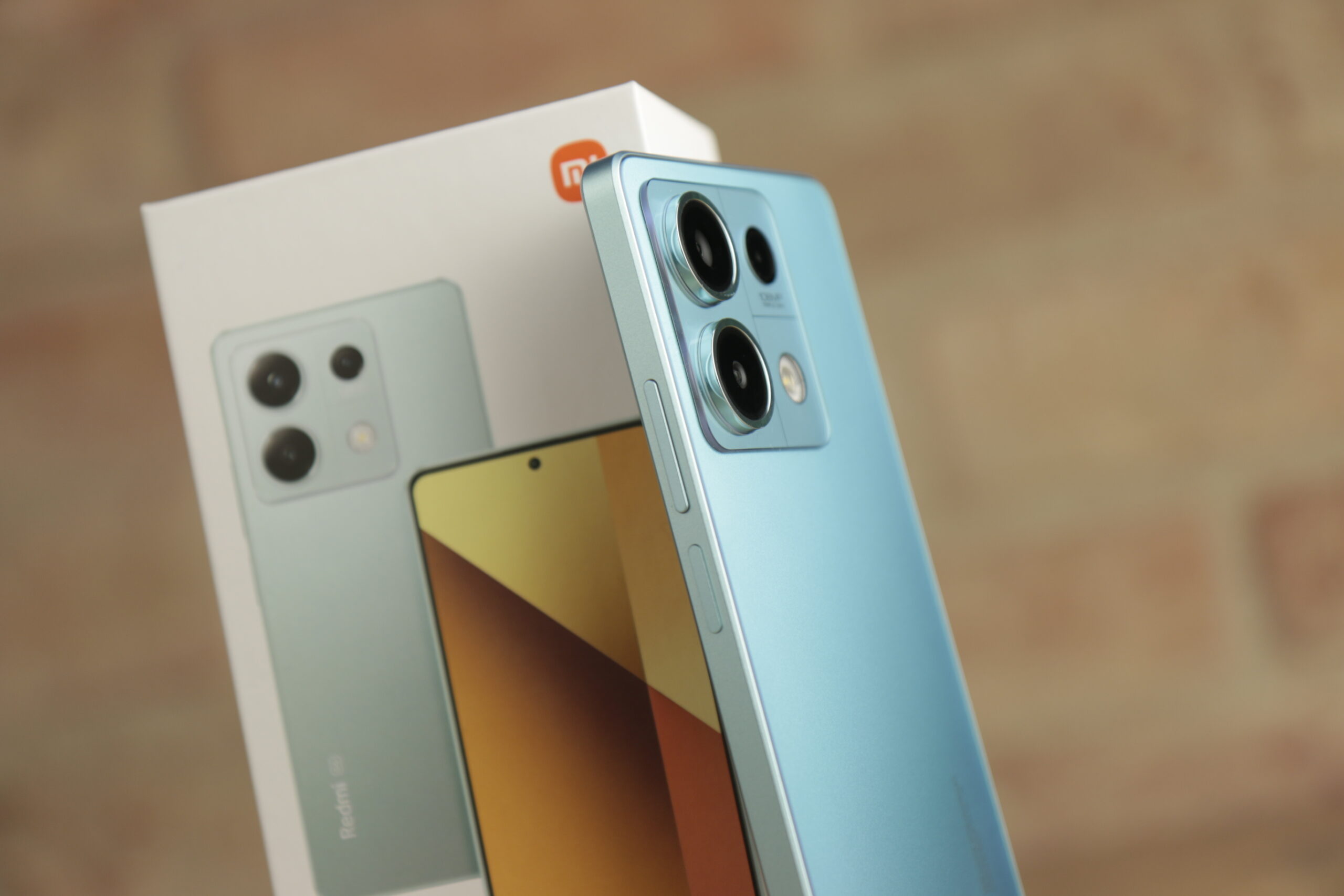 Testowany smartfon Redmi Note 13 5G koloru niebieskiego z potrójnym aparatem fotograficznym, oparty o pudełko z grafiką telefonu, na tle ceglanej ściany.