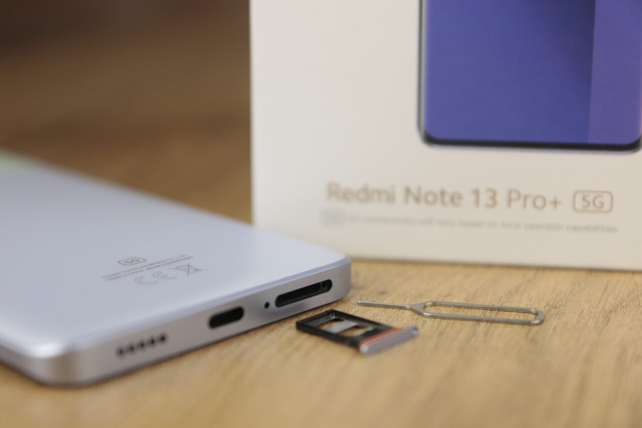 Białe tylne części smartfona obok pudełka z napisem "Redmi Note 13 Pro+ 5G", przy otwartym slocie na kartę SIM z założoną tacką i wyjętą igłą do wyjmowania tacy.