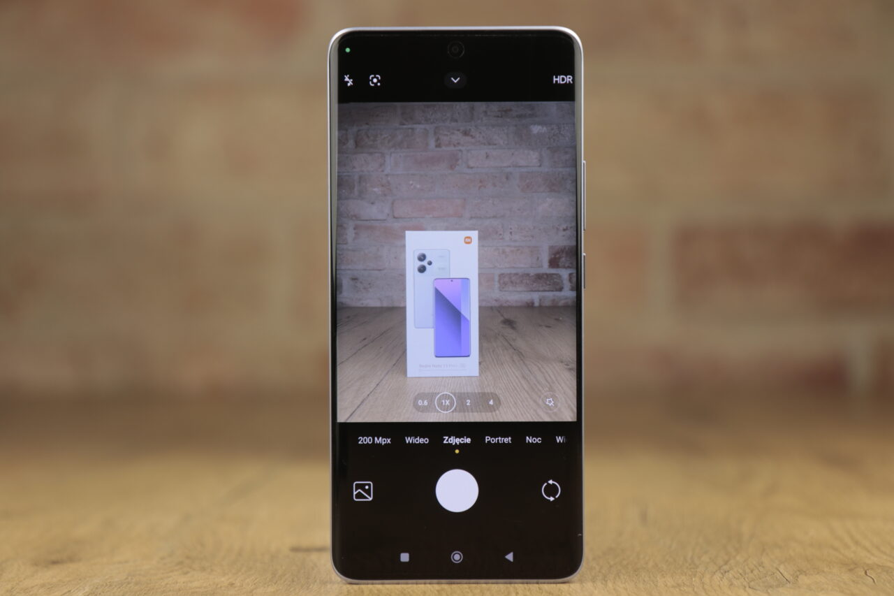 Recenzowany Redmi Note 13 Pro+ 5G z otwartą aplikacją aparatu skierowanego na pudełko z telefonem komórkowym, widocznym przez ekran smartfona, z tłem z ceglanej ściany.