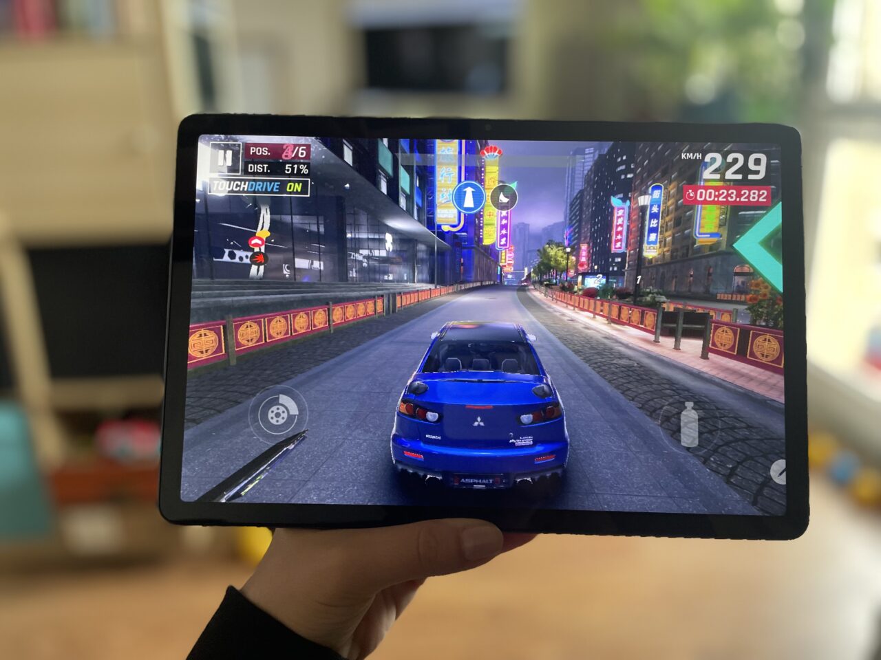 Ręka trzymająca tablet z wyświetlaną grą wyścigową, przedstawiającą niebieski samochód sportowy na miejskiej trasie w nocy.