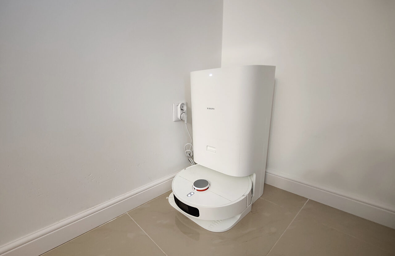 Um robô de limpeza branco da Xiaomi acoplado à estação de carregamento sob uma caldeira branca pendurada na parede.