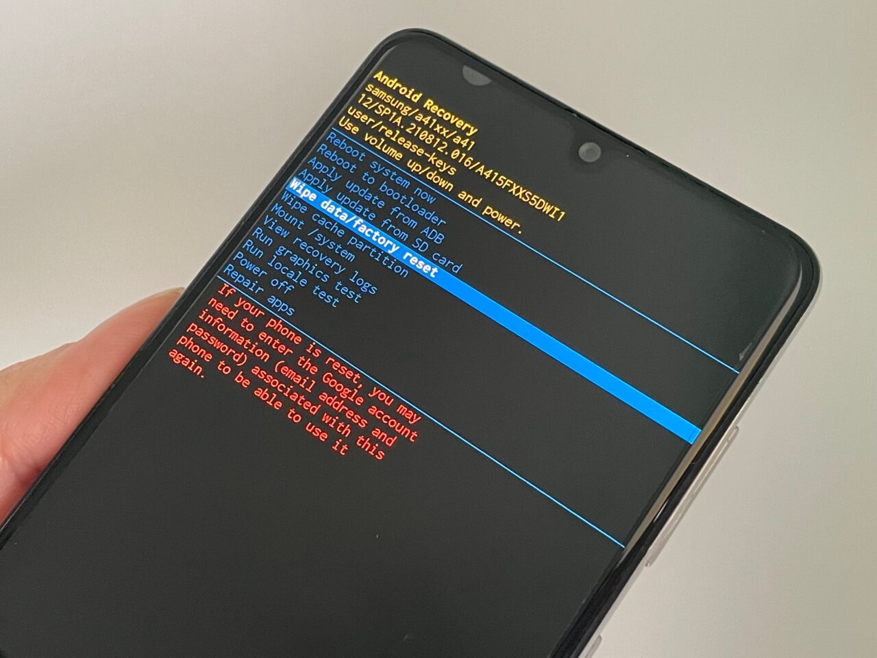 Smartfon trzymany w ręce, z włączonym ekranem pokazującym menu odzyskiwania systemu Android z różnymi opcjami i komunikatami w języku angielskim.