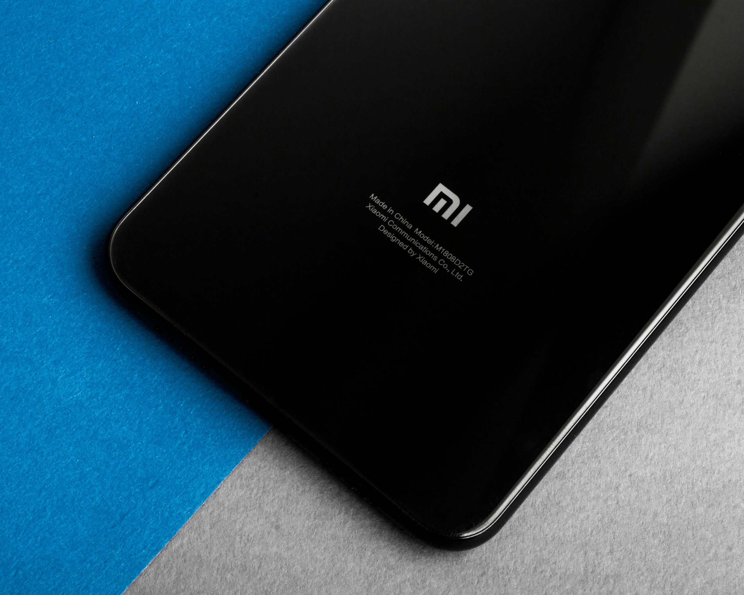 Częściowy widok czarnego smartfona Xiaomi z logo i napisami na tylnej obudowie, położonego na skos na dwóch powierzchniach - niebieskiej i szarej.
