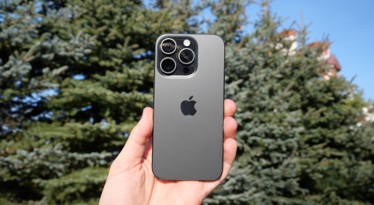 Smartfona Apple iPhone 15 Pro trzymany w ręku. W tle rozmazane drzewa iglaste i niebieskie niebo.