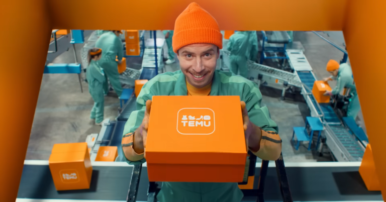 Mężczyzna w zielonym kombinezonie roboczym i pomarańczowej czapce trzyma pomarańczowe pudełko z logiem TEMU, w tle inni pracownicy sortujący paczki w centrum dystrybucji.
