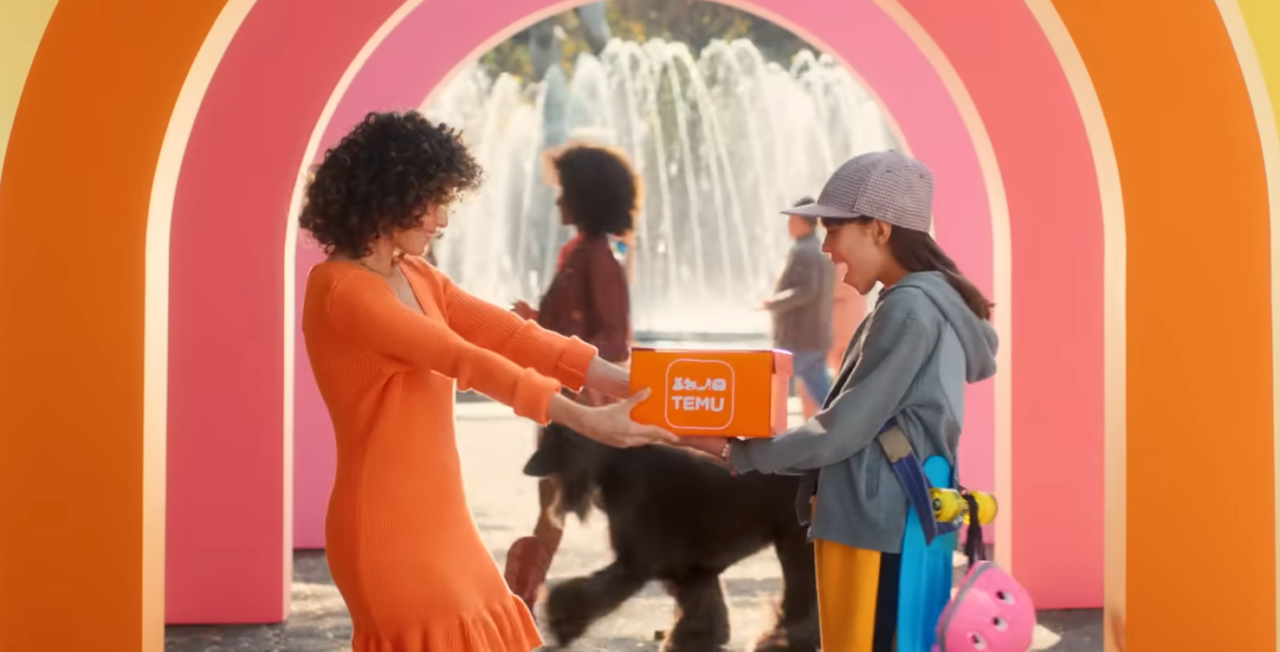 Dwie kobiety przekazują sobie kolorowe pudełko z napisem "TEMU" na tle fontanny i kolorowych, okrągłych bram.