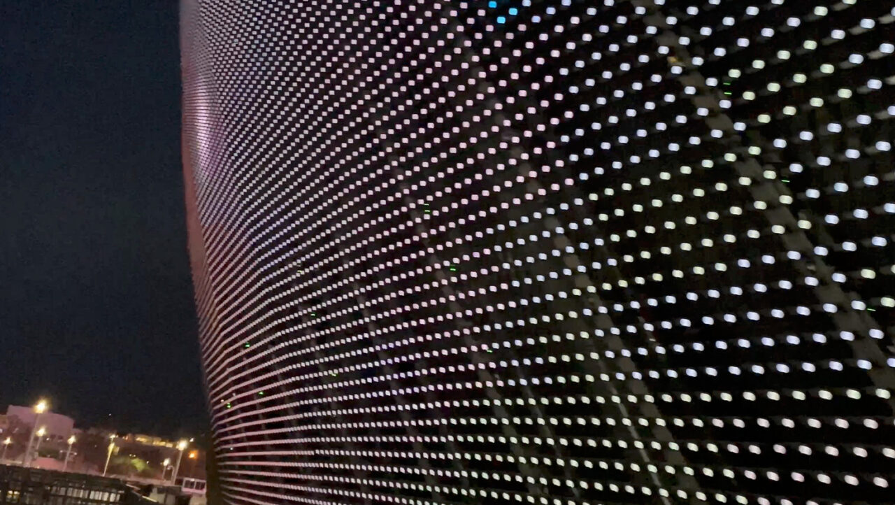 Zdjęcie pokazuje zbliżenie na zakrzywioną fasadę budynku pokrytą iluminacją LED tworzącą kropkowy wzór, z rozmytymi światłami miasta w tle.