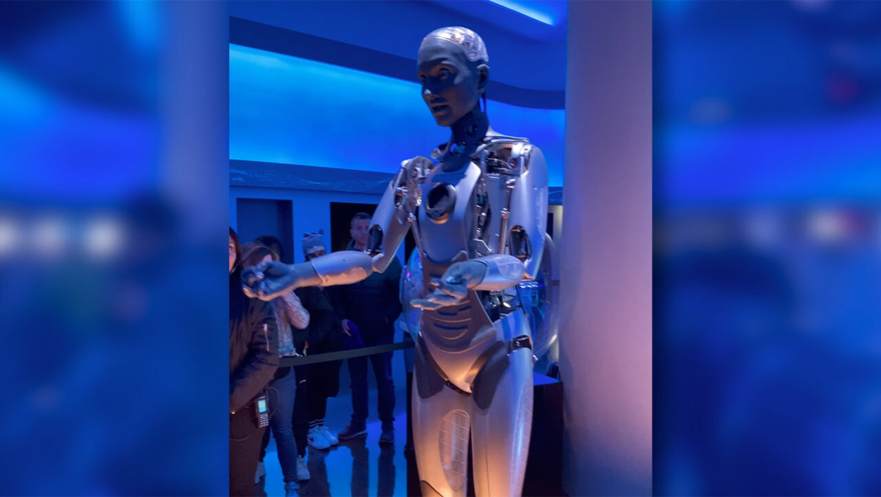 Robot humanoidzny z metalicznymi elementami na wystawie, na tle oświetlonego na niebiesko pomieszczenia z rozmytymi sylwetkami obserwujących ludzi.