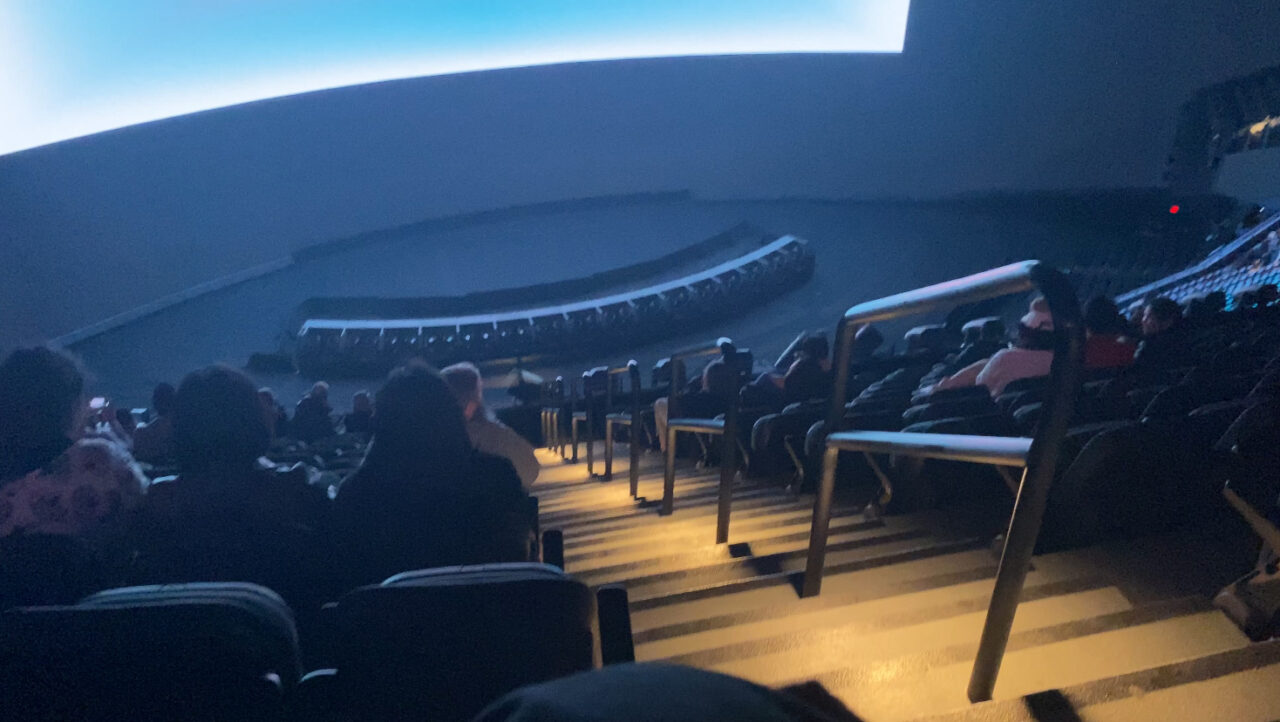 Widownia w półmroku z fotelami i publicznością, skupioną na wydarzeniu w planetarium z dużym, zakrzywionym ekranem.