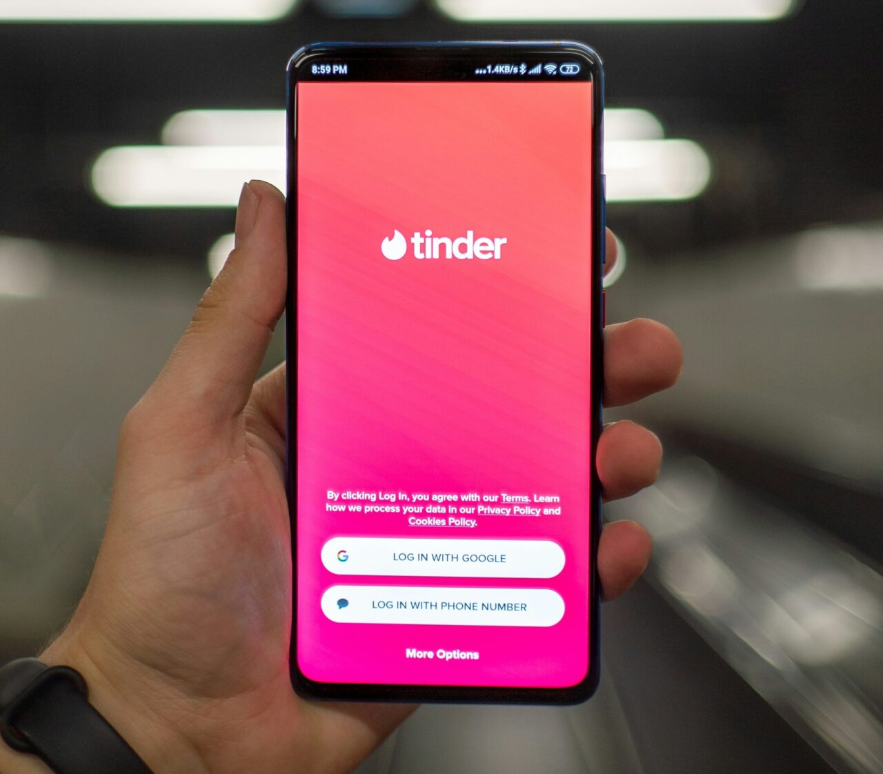 Osoba trzyma smartfon z ekranem logowania Tinder, programu idealnego na Walentynki z aplikacją randkową wyświetlającym opcje logowania przez Google i numer telefonu na tle rozmytych świateł.