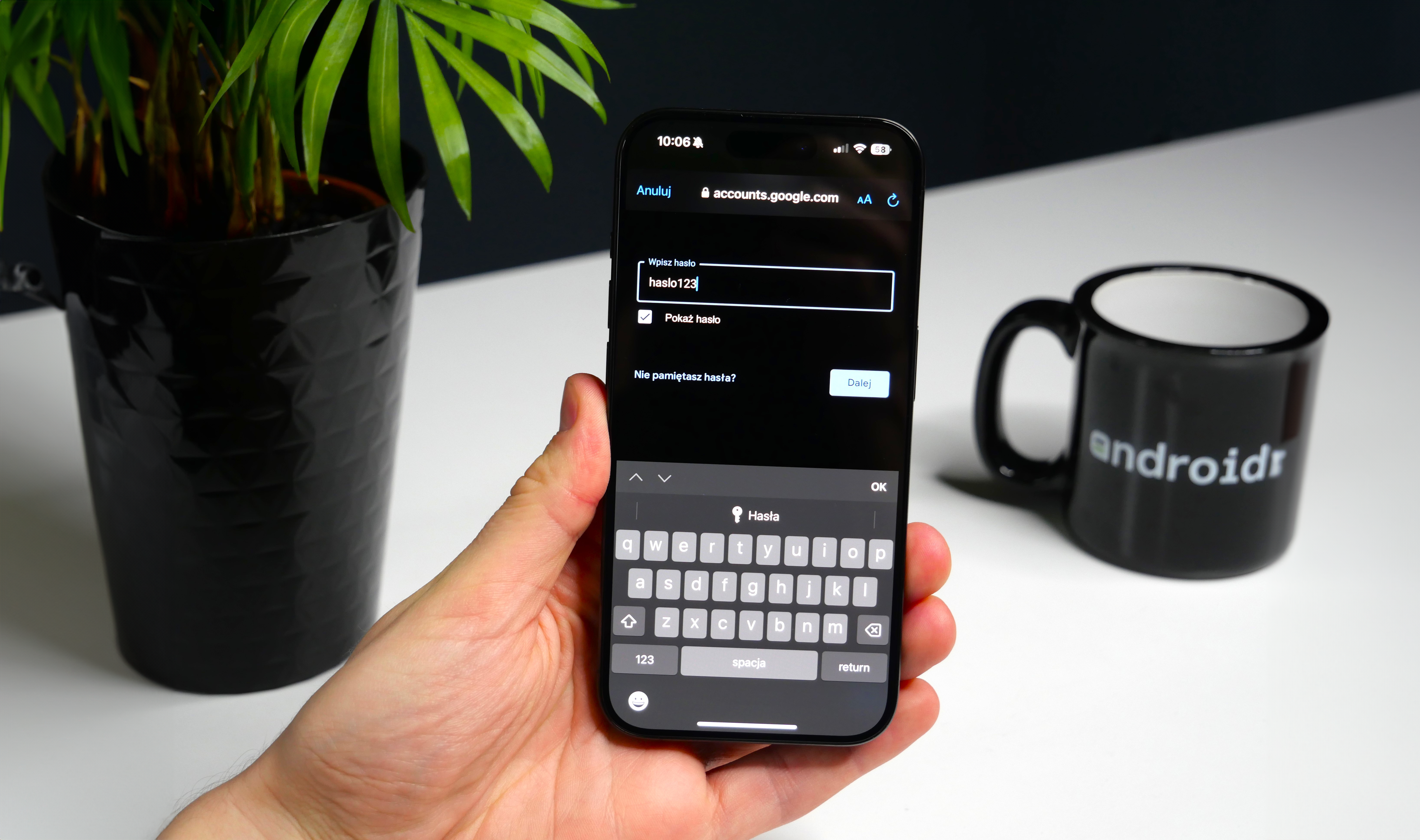 Ręka trzymająca smartfon z wyświetlaczem logowania do konta Google, na biurku doniczka z rośliną i kubek z napisem "android".