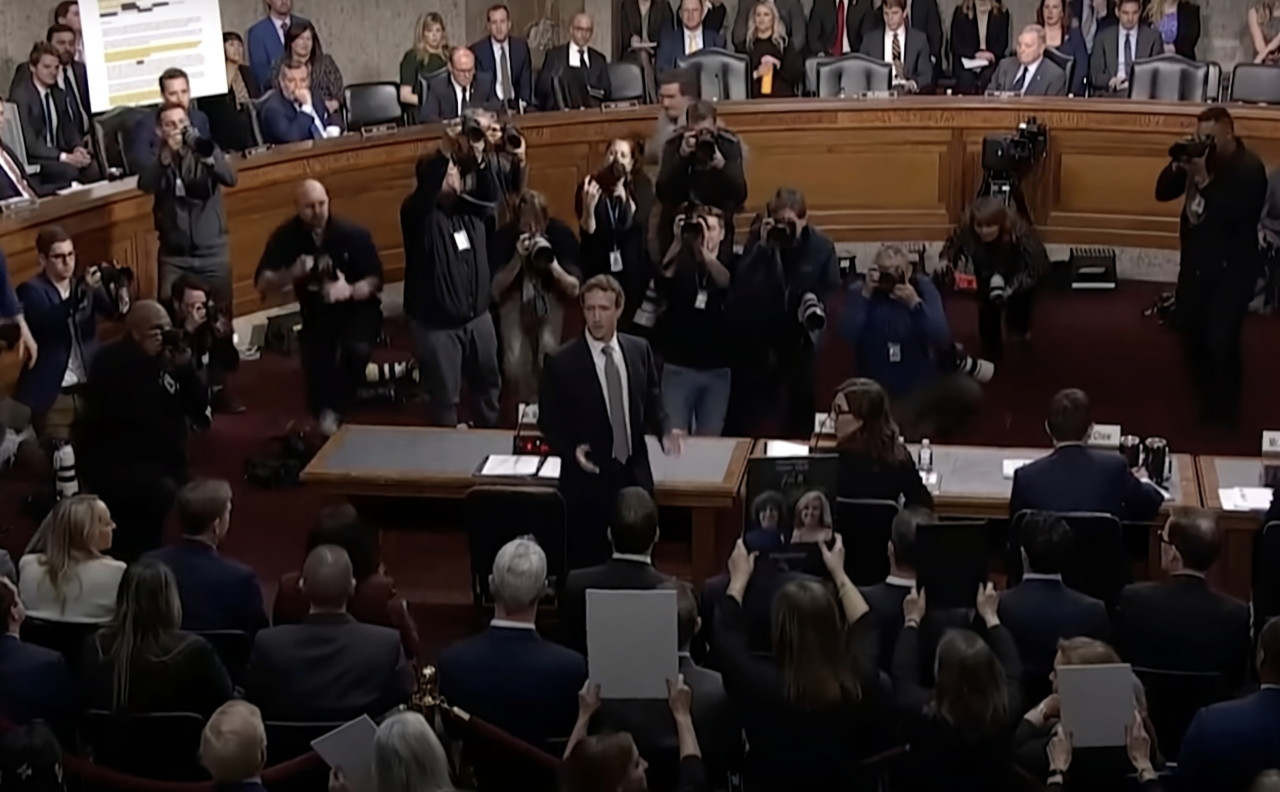 Sala sądowa pełna ludzi z Markiem Zuckerbergiem stojącym pośrodku i grupą fotografów robiących zdjęcia.