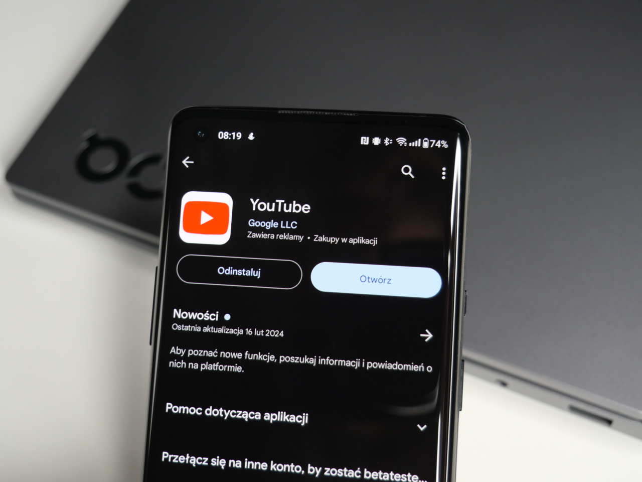 Smartfon wyświetlający informacje o aplikacji YouTube w sklepie z aplikacjami z przyciskami "Odinstaluj" i "Otwórz", z laptopem w tle.
