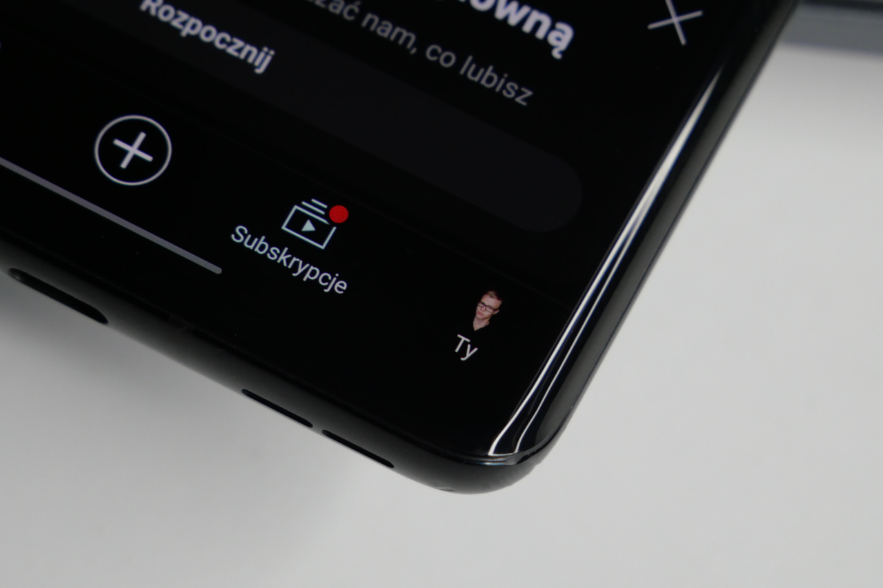 Smartfon leżący na białej powierzchni z wyświetlonym interfejsem aplikacji, pokazującym przycisk subskrypcji i miniaturę profilu użytkownika z napisem "Ty".