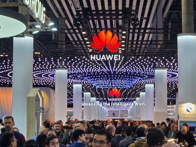 Tłum tłoczny na targach technologicznych z wyeksponowanym stoiskiem firmy Huawei, zdobionym ledowymi światłami i logotypami, w tle hasło "Advancing the Intelligent World".