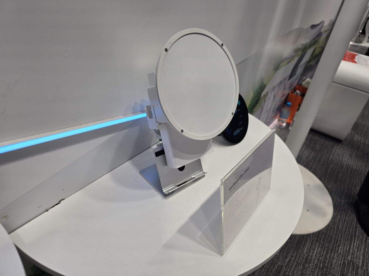 Biały, okrągły, modem 5G mmWave na stoisku Huawei 5.5G na targach MWC 2024. Biała jednostka z anteną mmWave w kształcie białego stożka na tle wystawowym, z niebieskim światłem LED i etykietą informacyjną.