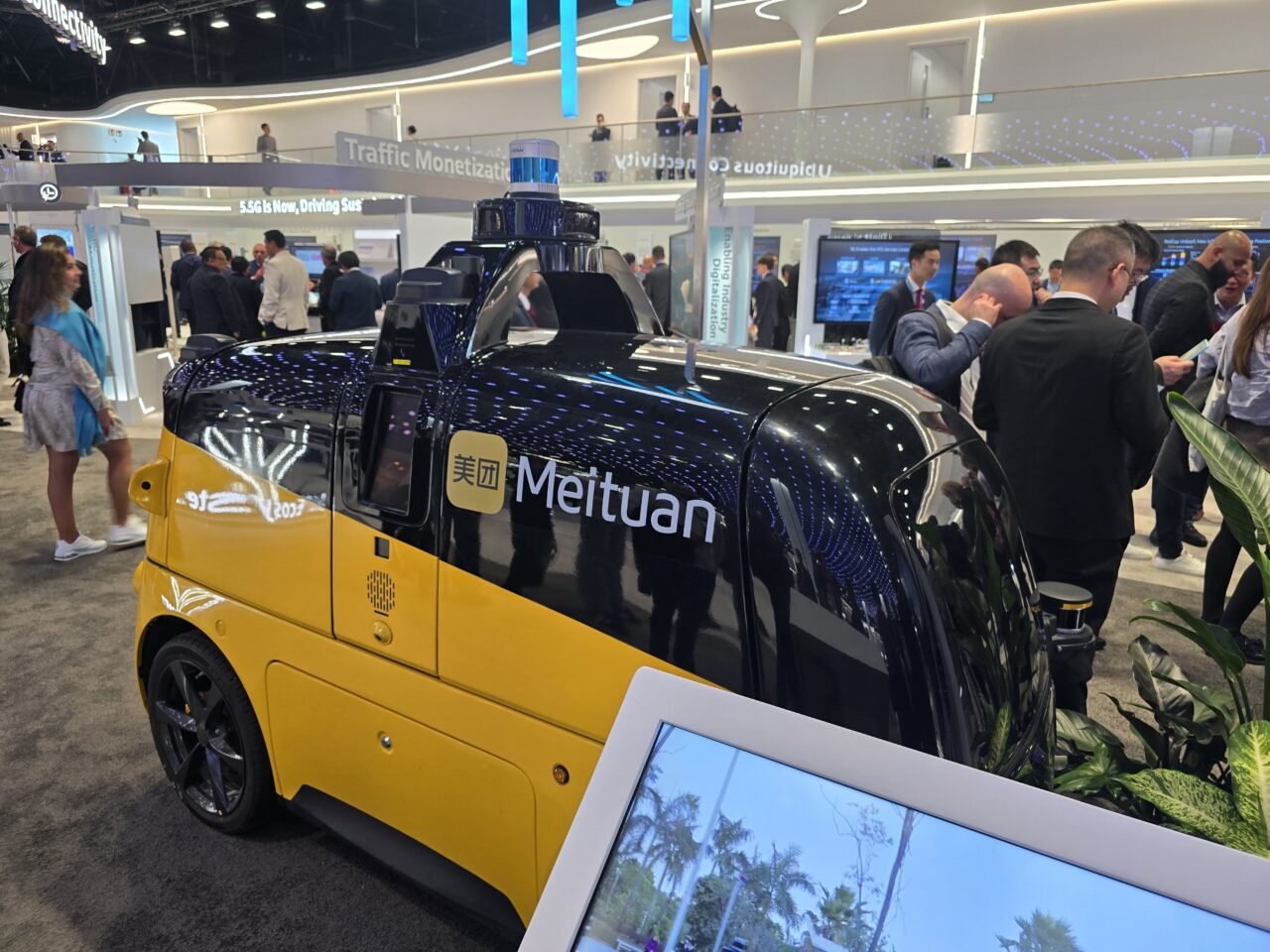 Samochód dostawczy Meituan na stoisku Huawei 5.5G na targach MWC 2024 z technologią autonomiczną na tle wystawy technologicznej, z ludźmi w tle rozmyślającymi o prezentowanych produktach.