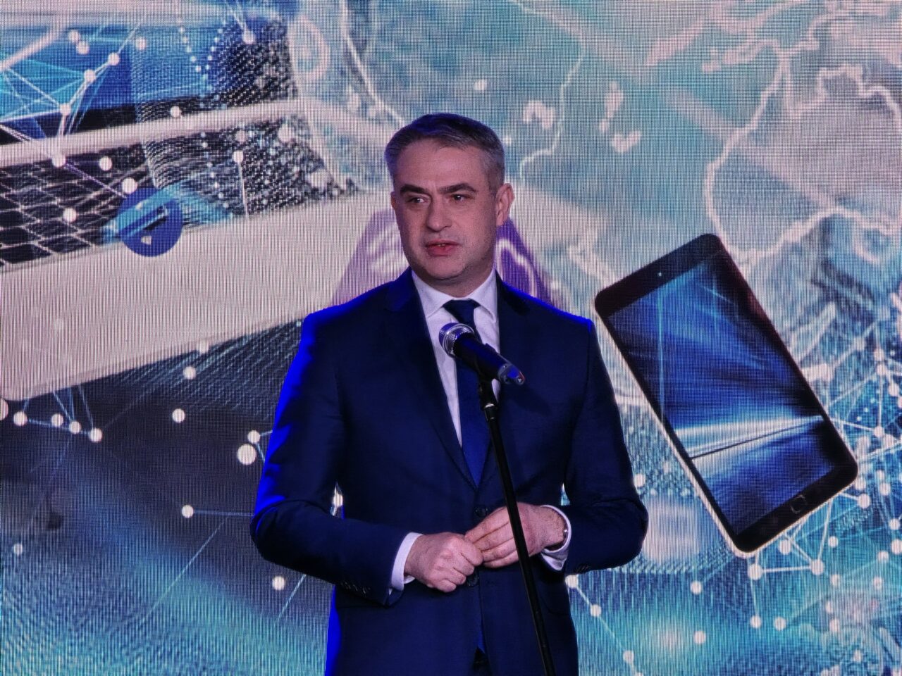 Minister cyfryzacji Krzysztof Gawkowski podczas 16. FG Time 2024. Mężczyzna w garniturze przemawiający przy mikrofonie na tle grafiki przedstawiającej sieci cyfrowe i unoszący się smartfon.