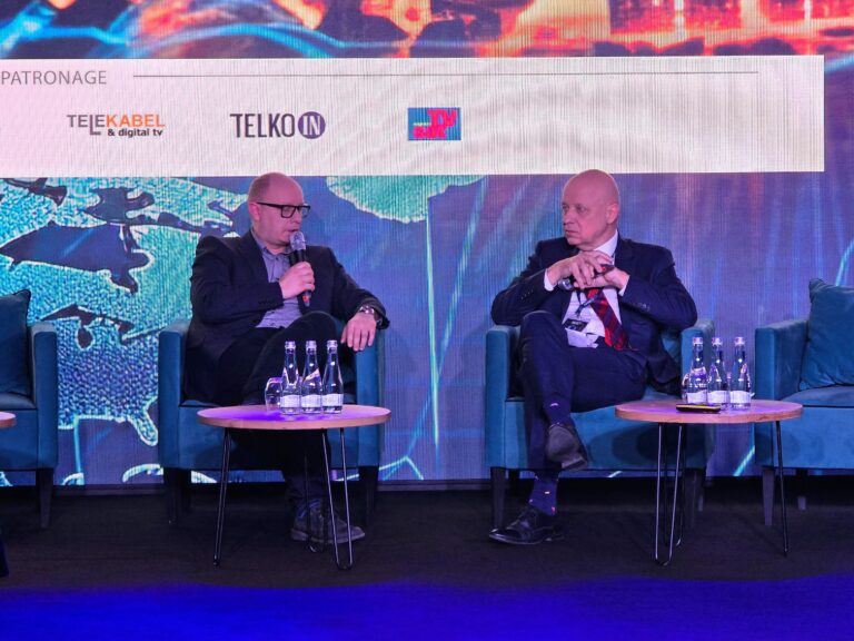 Dyskusja dot. AI w cyberbezpieczeństwie z udziałem dyrektora NASK (Radosław Nielek). Dwóch mężczyzn siedzi na scenie podczas wydarzenia branżowego, jeden z nich mówi do mikrofonu, w tle widoczne są logotypy sponsorów.