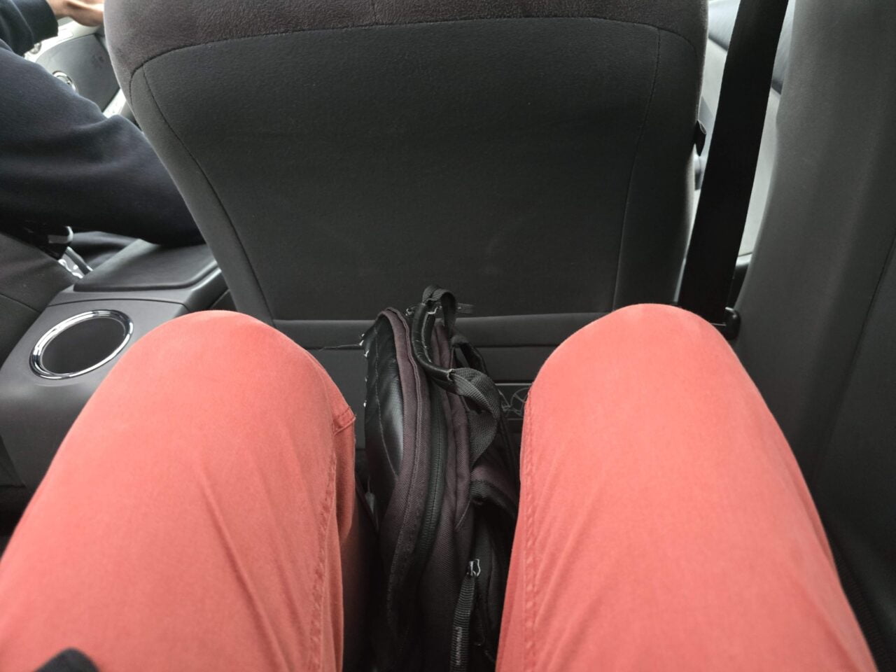 Osoba siedząca na tylnej kanapie samochodu, widok z góry na jej czerwone spodnie i plecak umieszczony między nogami.