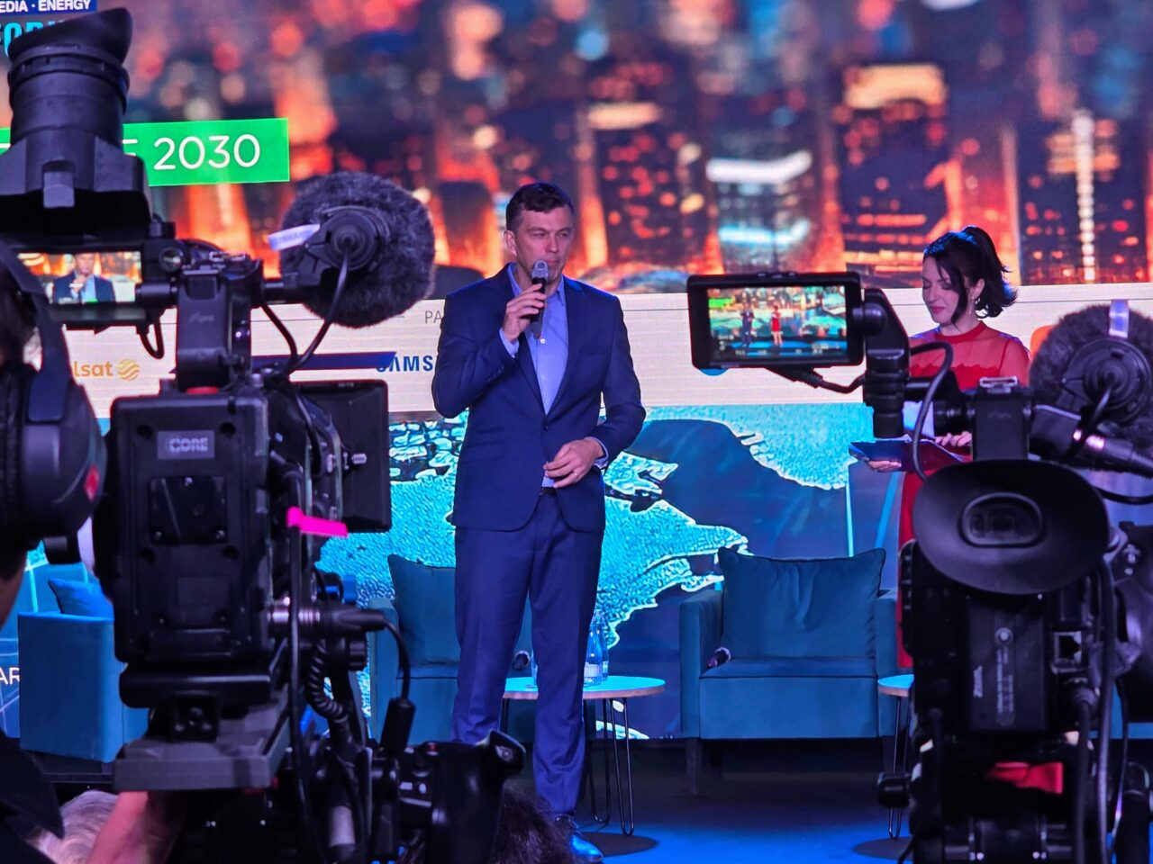 Drugi dzień 16. FG Time. Mężczyzna w garniturze przemawiający do mikrofonu na scenie przy dużej ekranie LED, kobieta w czerwonej sukni siedzi obok, widoczne kamery telewizyjne na pierwszym planie, tło sceny z miejską panoramą.