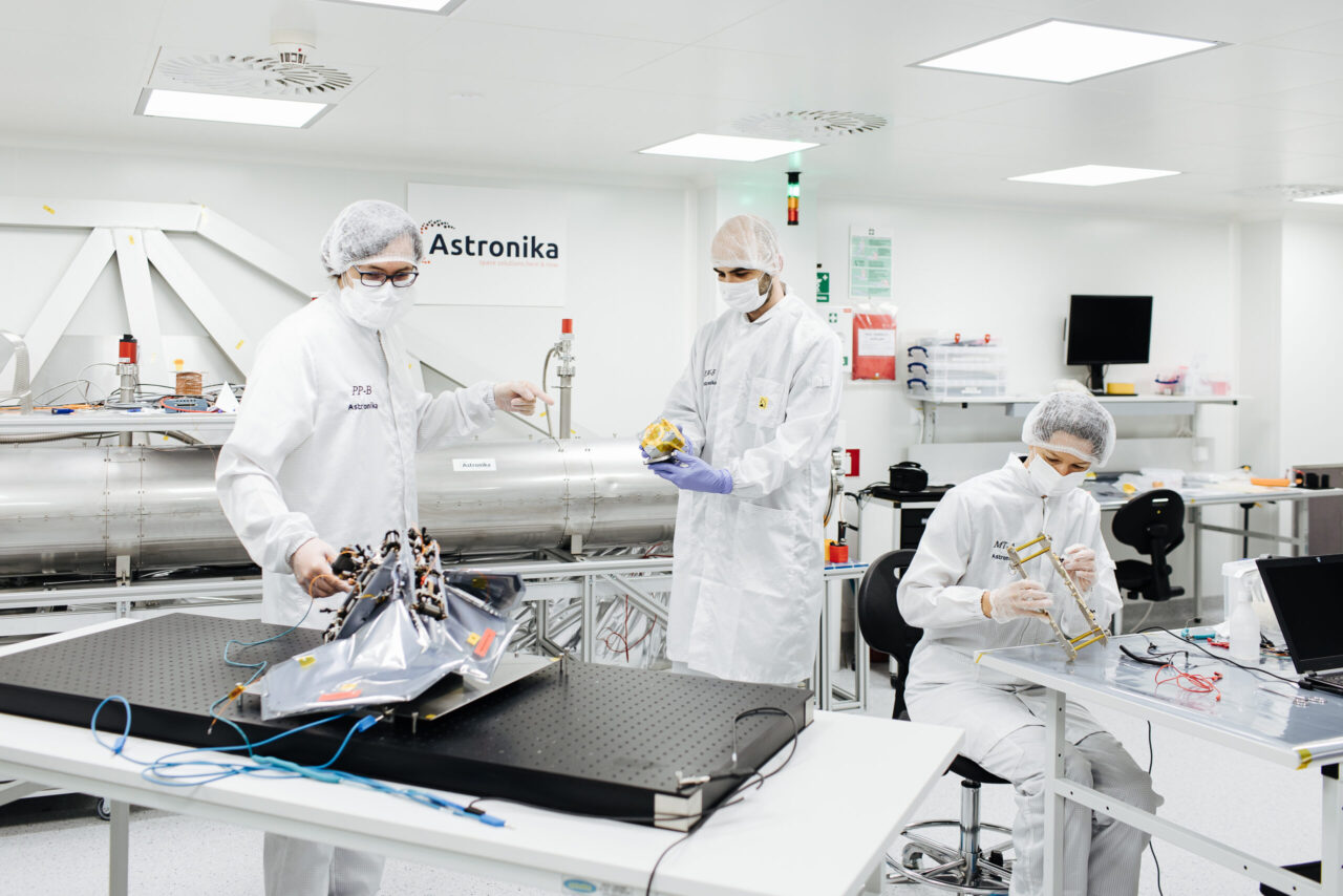 Trzy osoby w białych kombinezonach ochronnych pracują w laboratorium nad komponentami sprzętu kosmicznego.