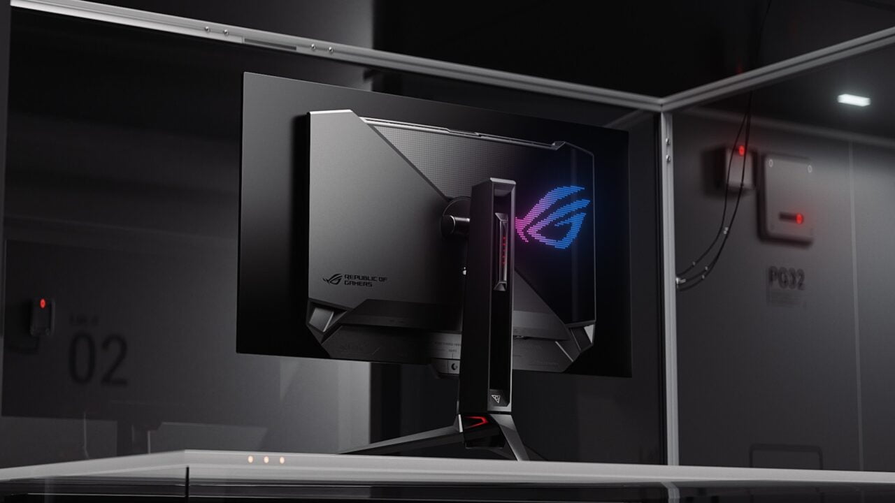 Monitor gamingowy Republic of Gamers umieszczony na biurku z widocznym podświetlanym logo i zaawansowanym systemem chłodzenia na tylnej obudowie, w ciemnym pomieszczeniu z kablami i wyłącznikami w tle.