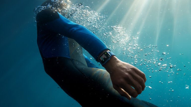 Nurek z zegarkiem inteligentnym na nadgarstku, zanurzony w wodzie z promieniami słonecznymi przenikającymi przez niebieską głębinę.