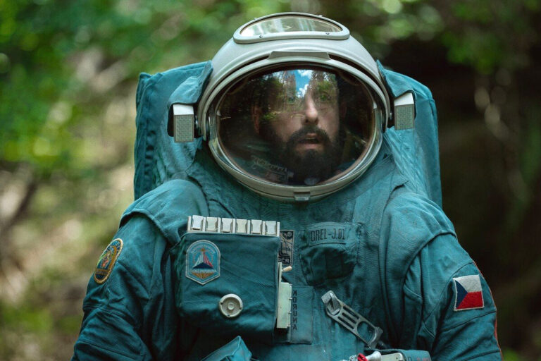 Adam Sandler jako Astronata w filmie Netflix. Mężczyzna w skafandrze astronauty w zielonym otoczeniu, z widocznym odbiciem drzew w jego hełmie.