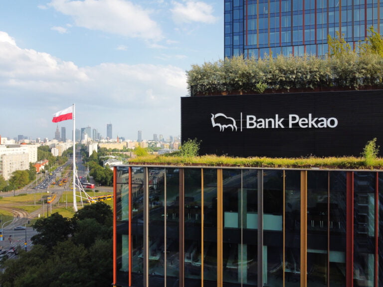 Widok na budynek Banku Pekao z zielonym dachem przed tłem miejskiej panoramy z polską flagą na maszcie.
