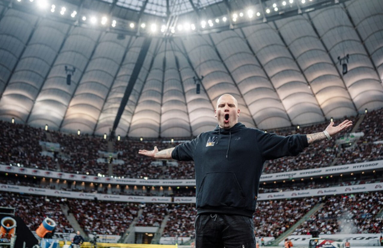 Budda w czarnej bluzie z rozłożonymi rękoma i otwartymi ustami stoi na pierwszym planie, w tle widoczni widzowie na trybunach zadaszonego stadionu.