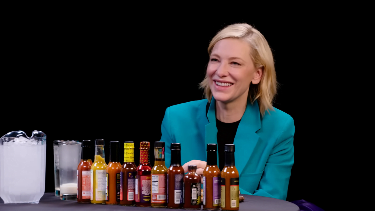 Cate Blanchett - kobieta o blond włosach i szerokim uśmiechu, ubrana w turkusowy żakiet, siedzi przy stole z różnorodnymi butelkami sosów pikantnych oraz szklanką mleka i kostką lodu.