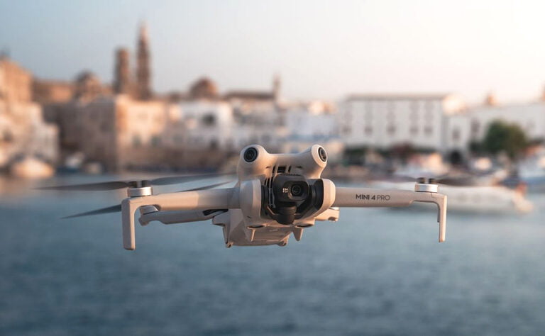 Dron DJI Mini 4 Pro Fly More Combo lecący na tle rozmytego miasta przy brzegu morza w złotym świetle zachodzącego słońca.