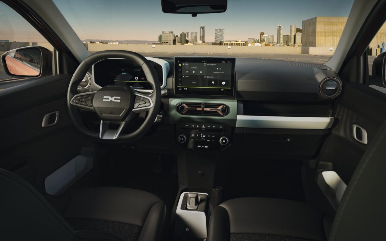 Widok z perspektywy kierowcy na wnętrze nowoczesnego samochodu ze skórzanym kierownicą, panelami z cyfrowymi wyświetlaczami, dużym ekranem dotykowym systemu multimedialnego w centralnej części deski rozdzielczej i automatyczną dźwignią zmiany biegów.