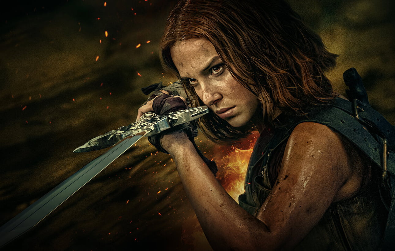 Kadr z filmu Dama od Netflix. Młoda kobieta o rudych włosach z zaciśniętą szczęką, trzymająca miecz, z tłem przedstawiającym unoszące się iskry i płomienie.