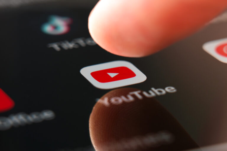 Palce dotykające ekran smartfona z wyeksponowaną ikoną aplikacji YouTube wśród innych nieostrych ikon aplikacji.