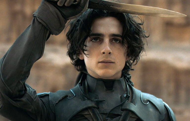 Kadr z filmu Diuna. Młody mężczyzna o ciemnych kręconych włosach, trzymający miecz tuż nad swoją głową w geście ochronnym, ubrany w ciemnozielony pancerz, na tle rozmytego beżowego otoczenia.